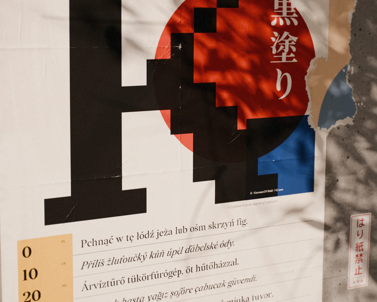A poster set in Kuronuri on a wall, 黒塗り over a pixelated red sun, above pangrams in Polish, Czech, and Hungarian